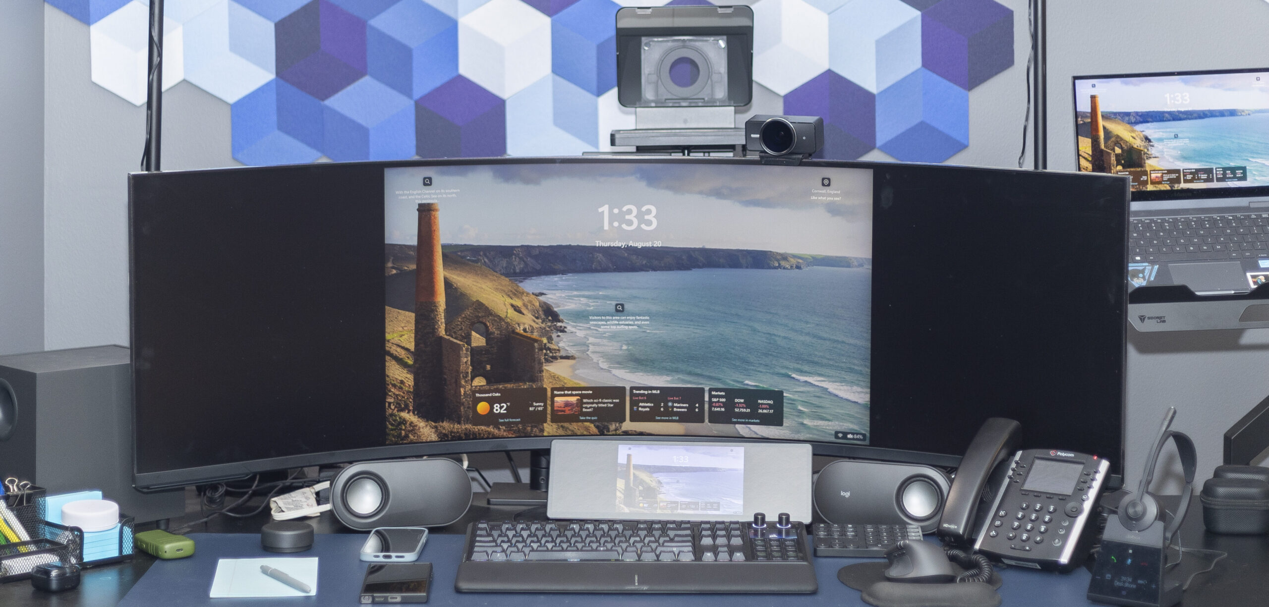 A home sales desk seen across the desktop: curved ultrawide monitor, keyboard, mouse, speakers and a laptop on a monitor arm.