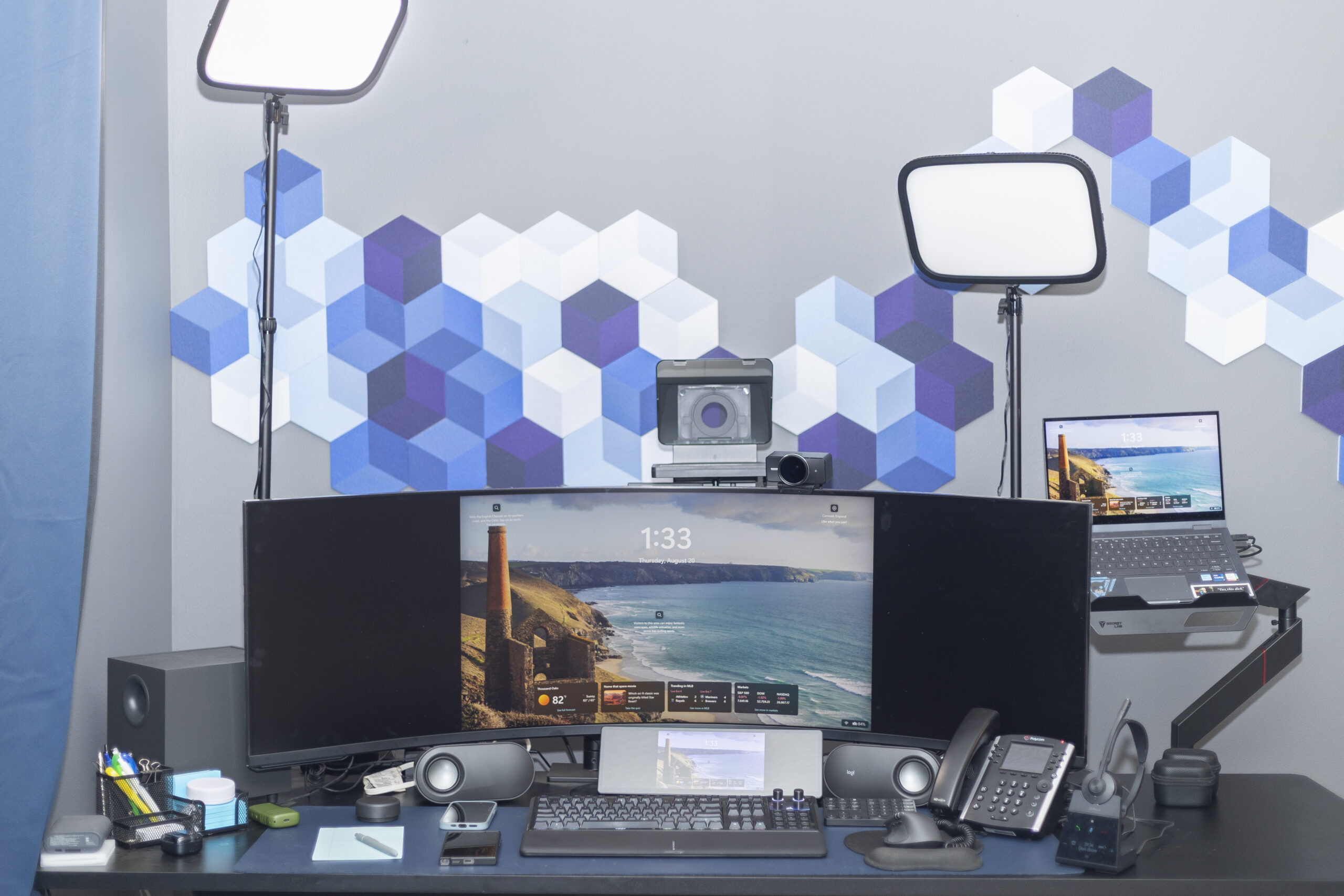 A home sales desk: a curved ultrawide monitor, split keyboard, laptop on a monitor arm, desk phone, wireless headset, speakers and two key lights against an acoustic-panelled wall.