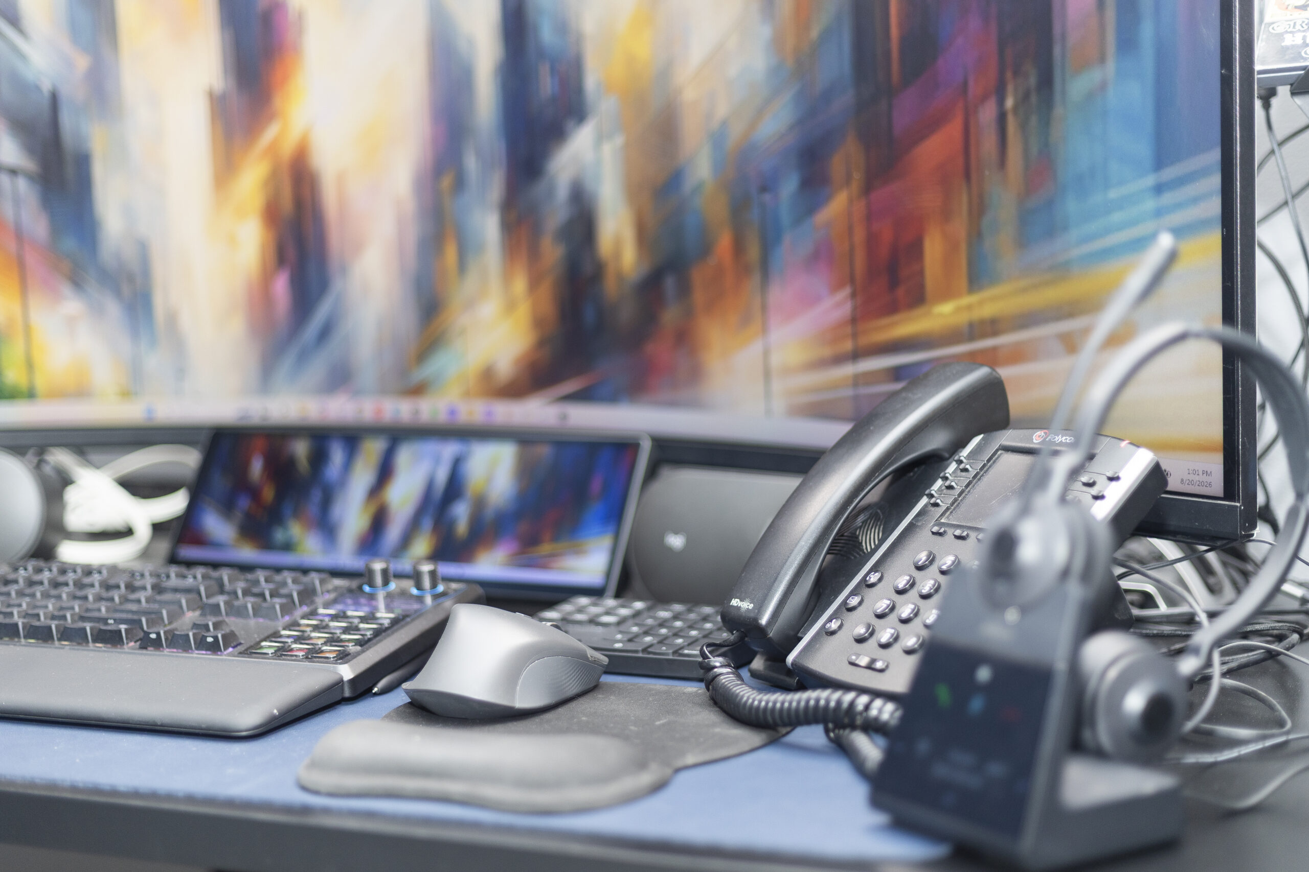 A close view of a home sales desk: a Polycom desk phone with the handset in its cradle, a wireless headset on its base, an ergonomic mouse and keyboard, and an ultrawide monitor behind.