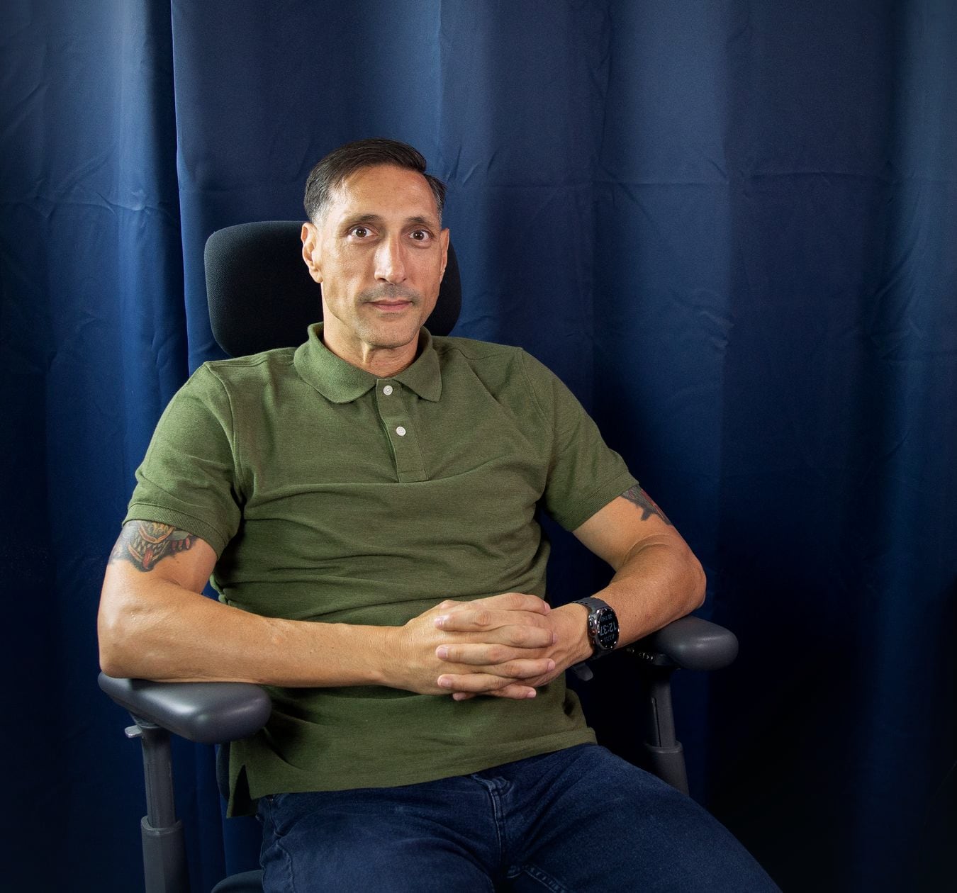 Jarrod Roberts sitting back in his office chair against a navy curtain, hands clasped, looking straight at the camera.
