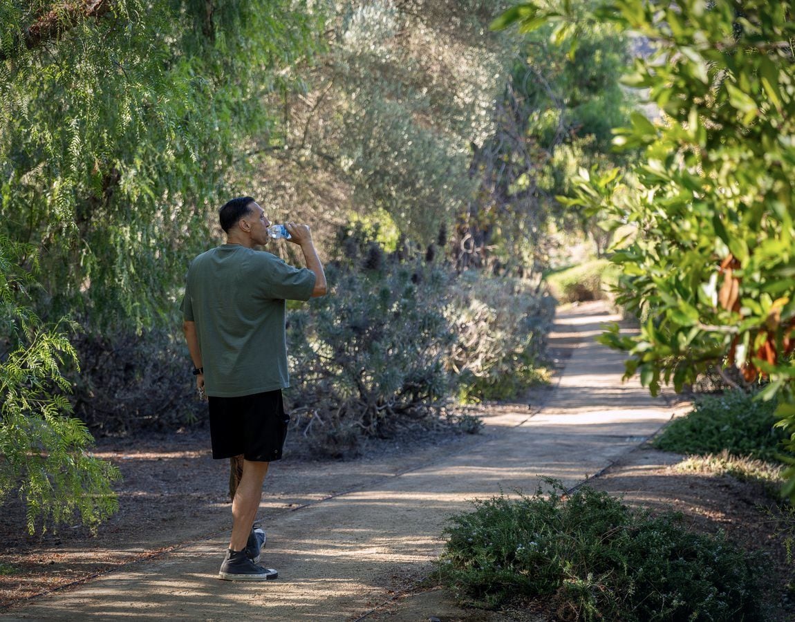 Jarrod Roberts on a morning walk along a shaded garden path, drinking from a water bottle mid-stride.